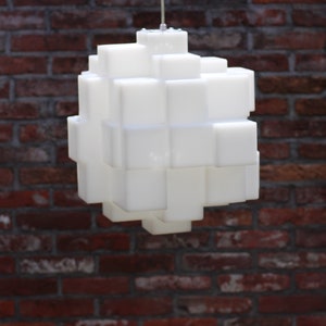 Op de afbeelding: Een witte, kubusvormige hanglamp hangt aan het plafond. De lamp is gemaakt van veel kleine, witte kubussen die aan elkaar zijn verbonden.