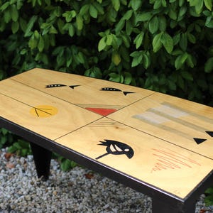 Peut inclure: Table basse rectangulaire avec un dessus en bois clair orné de motifs géométriques abstraits noirs, rouges et jaunes. La table a des pieds noirs et un bord noir. La table mesure environ 122 cm de long.
