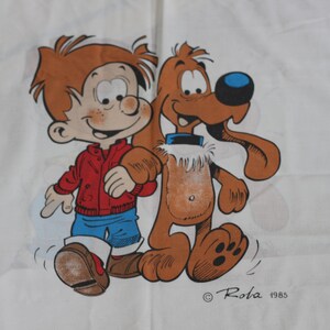 May include: A white tea towel with a blue border features a cartoon illustration of a boy with red hair and a red jacket walking with a brown dog. The text "Rola 1985" is printed in the bottom right corner.