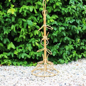 Può includere: Un portapiante alto in bambù naturale con più supporti circolari e a forma di gancio. Il supporto ha una parte superiore decorativa e una base circolare ampia. È ambientato su uno sfondo di fogliame verde e piccoli ciottoli bianchi.