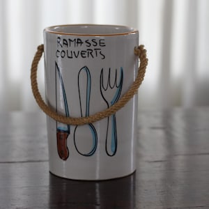 Puede incluir: Un porta cubiertos de cerámica blanca con un asa de cuerda. El porta cubiertos tiene una ilustración azul y negra de un cuchillo, un tenedor y una cuchara con el texto "Ramasse couverts" en la parte delantera.
