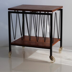 Puede incluir: Un revistero rodante de estilo moderno de mediados de siglo con una tapa de madera y un marco de metal negro. El revistero tiene una serie de barras de metal curvadas para sujetar las revistas y est&aacute; sobre cuatro ruedas.