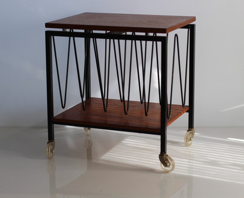 Puede incluir: Un revistero rodante de estilo moderno de mediados de siglo con una tapa de madera y un marco de metal negro. El revistero tiene una serie de barras de metal curvadas para sujetar las revistas y est&aacute; sobre cuatro ruedas.