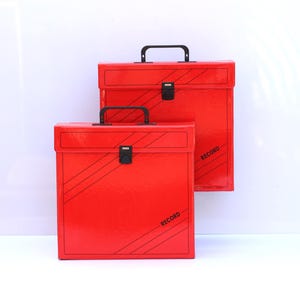 Puede incluir: Dos cajas rojas para guardar discos con asas y cierres negros. Las cajas tienen líneas diagonales negras y la palabra "RECORD" impresa en el lateral. Forma rectangular y acabado brillante.