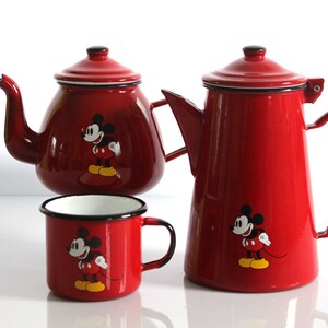 May include: A red enamelware tea set featuring Mickey Mouse. The set includes a teapot, a coffee pot, and a mug. Each piece has a black rim and handle, and a Mickey Mouse graphic. The teapot and coffee pot have lids.