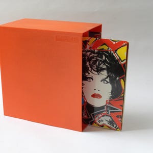 May include: A bright orange rectangular slipcase partially housing a book or box set. The visible portion of the inner item features a graphic illustration of a person with dark hair, red lips, and bold yellow and red geometric patterns.