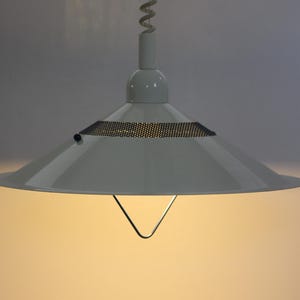 May include: White pendant light with a conical shade and a mesh detail. The light has a simple, modern design, with a metal frame and a wire hanging from the ceiling. The light is on, casting a warm glow.