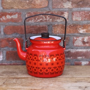 May include: A red enamel teapot with a black handle and lid. The teapot features a floral pattern in shades of orange and red. The teapot is sitting on a wooden surface in front of a brick wall.