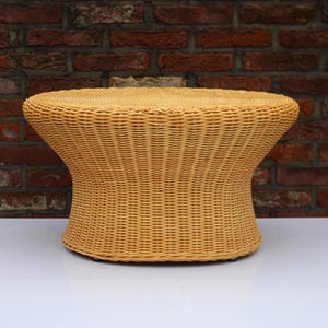 May include: A light brown wicker side table with a round top and a curved base. The table is made of woven wicker and is set against a brick wall.