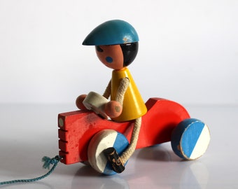 May include: A vintage wooden pull toy featuring a figure in a blue cap and yellow shirt riding a red tricycle. The wheels are painted with blue and white accents, and a blue string is attached for pulling.