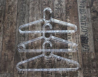 May include: Three clear plastic clothes hangers stacked vertically. Each hanger features a triangular frame with a central hook at the top and small notches along the horizontal bar for securing garments. The material is transparent and smooth.