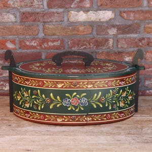 Peut inclure: Boîte ovale en bois peinte à la main avec un couvercle. La boîte est principalement vert foncé avec des motifs floraux et de feuilles rouges, blancs et bleus. Elle a une poignée et des poignées latérales marron foncé. Fond en briques.