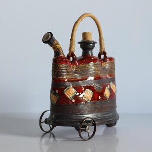 Op de afbeelding: Een rode, bruine en gouden keramische theepot met een kurkstop en een geweven handvat. De theepot heeft een geometrisch patroon en staat op wielen.