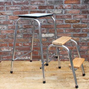 May include: A chrome metal step stool featuring a tall frame with a white top seat and two lower steps made of light-colored wood. The structure is supported by four tubular legs with black rubber feet, providing a stable base for reaching high areas.