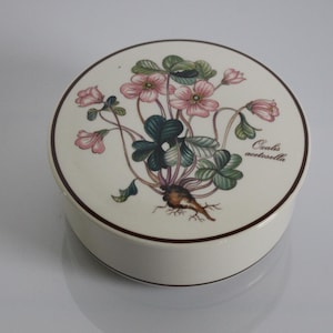 Op de afbeelding: Ronde, witte keramische sieradendoos met een bloemenontwerp. Het deksel heeft een botanische illustratie van roze bloemen, groene bladeren en de tekst "Oxalis acetosella". De doos heeft een donkerbruine rand.