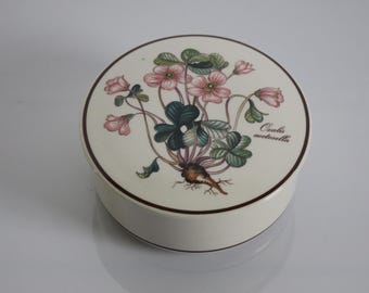 Może przedstawiać: Okrągłe, białe ceramiczne pudełko na drobiazgi z kwiatowym wzorem. Pokrywka przedstawia botaniczną ilustrację różowych kwiatów, zielonych liści i napis "Oxalis acetosella". Pudełko ma ciemnobrązową obwódkę.