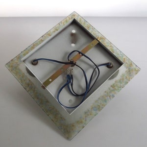 May include: The back of a square, white ceiling light fixture with a decorative, mosaic-like border. The fixture has three electrical wires and a metal bracket.
