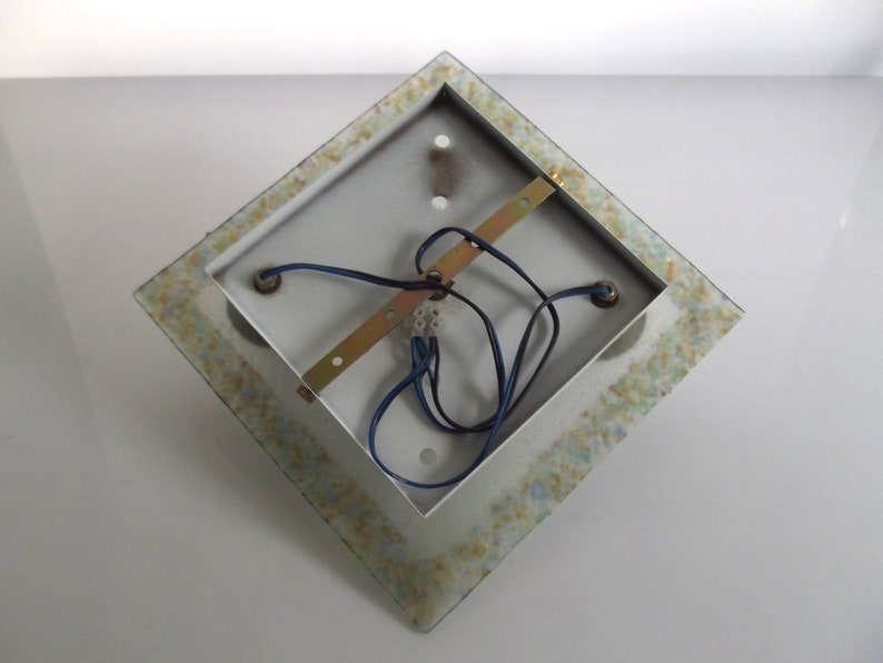 May include: The back of a square, white ceiling light fixture with a decorative, mosaic-like border. The fixture has three electrical wires and a metal bracket.
