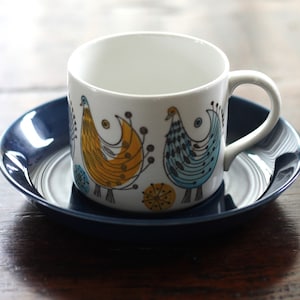 Op de afbeelding: Een witte theekop van keramiek met een blauw en geel vogelontwerp staat op een donkerblauwe schotel. De kop heeft een handvat en de schotel heeft een lichte rand.