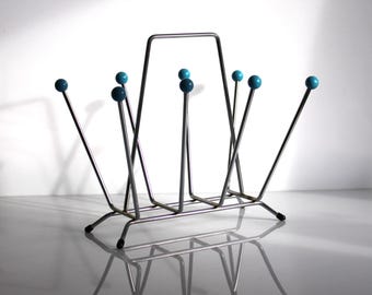 Peut inclure: Un porte-revues chromé avec des accents de boules turquoise. Le porte-revues a une base rectangulaire et une poignée en haut. Les boules turquoise sont aux extrémités de fines tiges métalliques.