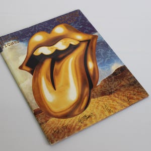 Op de afbeelding: Een vintage Rolling Stones albumhoes met het iconische gouden tong- en lippenlogo. De illustratie toont een landschap met een blauwe lucht en bruine heuvels. Het woord "STONE" is zichtbaar.