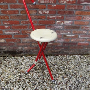 May include: A red metal folding stool with a white round seat and a curved back. The stool is standing on a gravel surface.