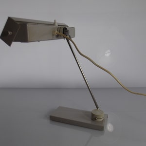 K&ouml;nnte beinhalten: Eine silberne Schreibtischlampe mit einem langen, verstellbaren Arm und einem rechteckigen Sockel. Die Lampe hat ein wei&szlig;es Kabel und einen wei&szlig;en Schalter.