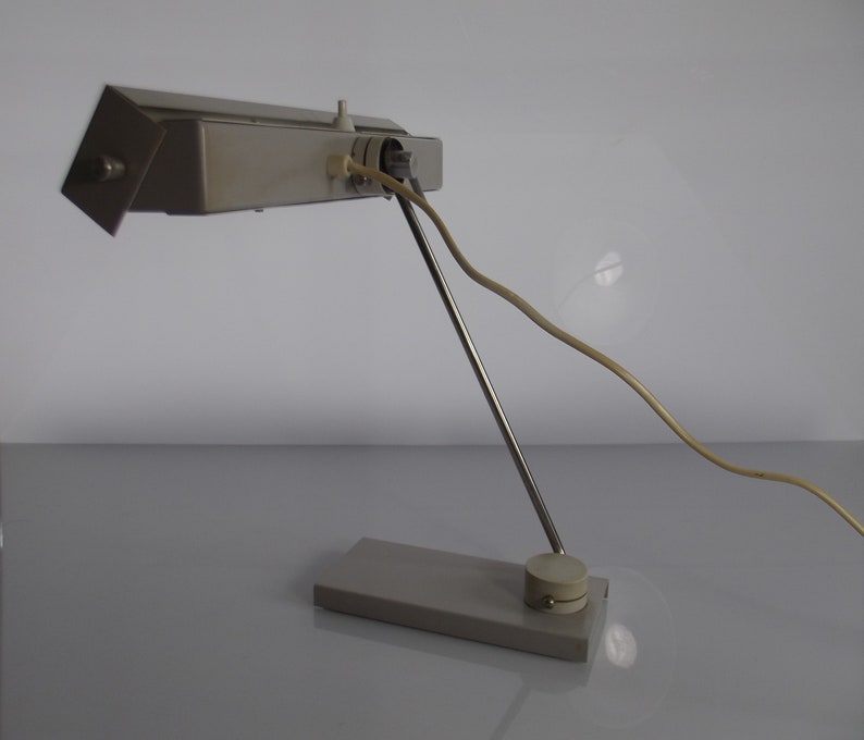 K&ouml;nnte beinhalten: Eine silberne Schreibtischlampe mit einem langen, verstellbaren Arm und einem rechteckigen Sockel. Die Lampe hat ein wei&szlig;es Kabel und einen wei&szlig;en Schalter.