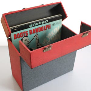 Puede incluir: Una caja de almacenamiento de discos de vinilo roja y gris vintage con tapa con bisagras. La caja está abierta, revelando un álbum con el texto "BOOTS RANDOLPH" y un hombre tocando el saxofón. La caja tiene un asa y cierres metálicos.