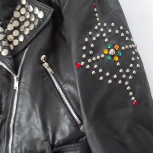 May include: Black leather jacket with silver studs on the collar and a decorative pattern on the sleeve. The sleeve design includes colorful gemstones. The jacket has a zipper closure and pockets.