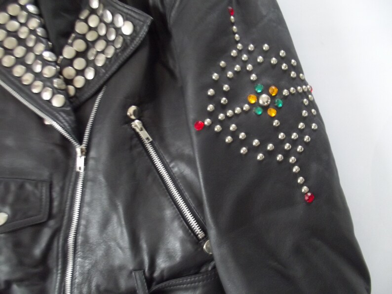 May include: Black leather jacket with silver studs on the collar and a decorative pattern on the sleeve. The sleeve design includes colorful gemstones. The jacket has a zipper closure and pockets.