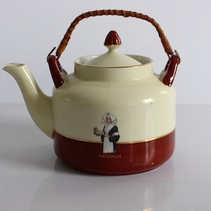 May include: Cream-colored ceramic teapot with a burgundy base and accents. Features a woven handle and a lid with a burgundy finial. A small illustration of a woman in a habit is on the front, with the text "CACAO CHOCOLATE".