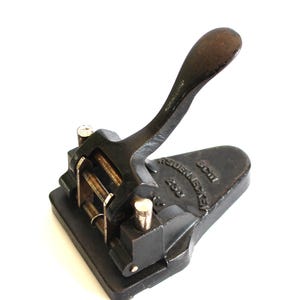 Op de afbeelding: Een vintage zwarte metalen perforator met een hendel. De perforator heeft een rechthoekige basis en een gebogen handvat. De tekst "Veydenneker 200" is zichtbaar aan de zijkant. Ontworpen om gaten in papier te maken.