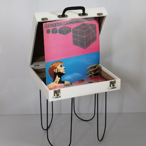Op de afbeelding: Een witte platenspelerstandaard met zwarte haarspeldpoten. De standaard is open en toont een vinylplaat met een roze en blauwe hoes. De plaat heeft de tekst "BERLIN BLONDES" en geometrische vormen. De standaard heeft een zwarte handgreep.