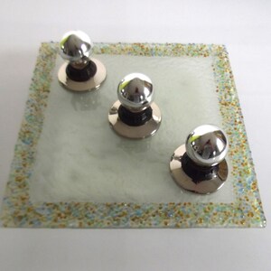 May include: Three silver knob pulls with round, shiny tops, set on a square, clear glass tray with a speckled border.