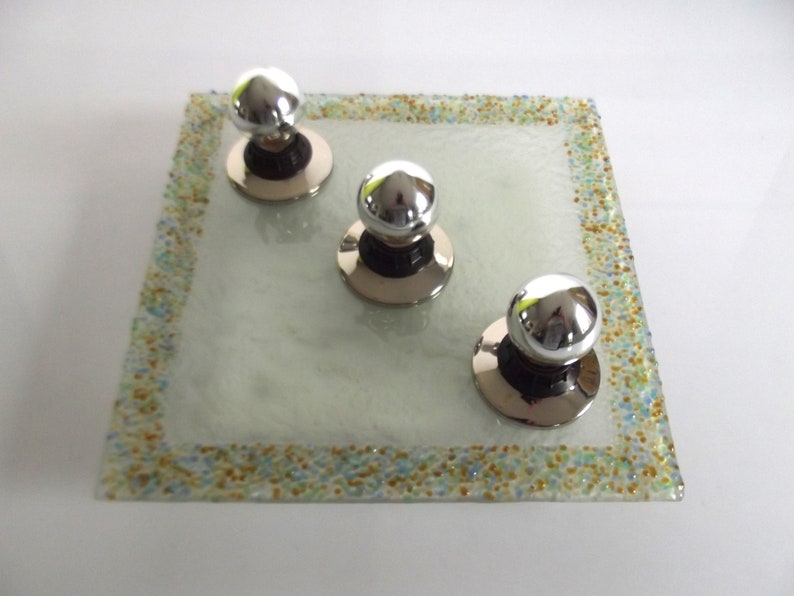 May include: Three silver knob pulls with round, shiny tops, set on a square, clear glass tray with a speckled border.