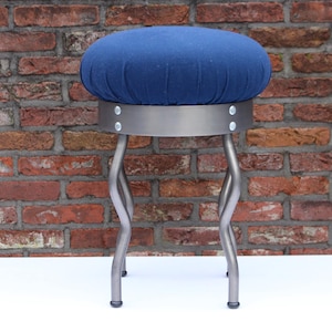 May include: A blue upholstered stool with a round seat and a metal base. The seat is a deep blue color, and the metal base has a silver finish. The legs are curved and the stool is set against a brick wall.