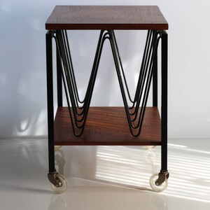 Puede incluir: Una mesa auxiliar de estilo moderno de mediados de siglo con una tapa de madera oscura y un marco de metal negro. La mesa presenta un dise&ntilde;o &uacute;nico con una serie de varillas met&aacute;licas curvadas que crean un patr&oacute;n decorativo. La mesa est&aacute; sobre ruedas y tiene un estante inferior.