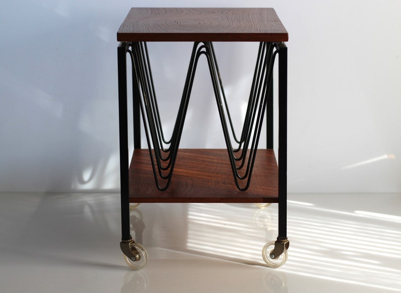 Puede incluir: Una mesa auxiliar de estilo moderno de mediados de siglo con una tapa de madera oscura y un marco de metal negro. La mesa presenta un dise&ntilde;o &uacute;nico con una serie de varillas met&aacute;licas curvadas que crean un patr&oacute;n decorativo. La mesa est&aacute; sobre ruedas y tiene un estante inferior.