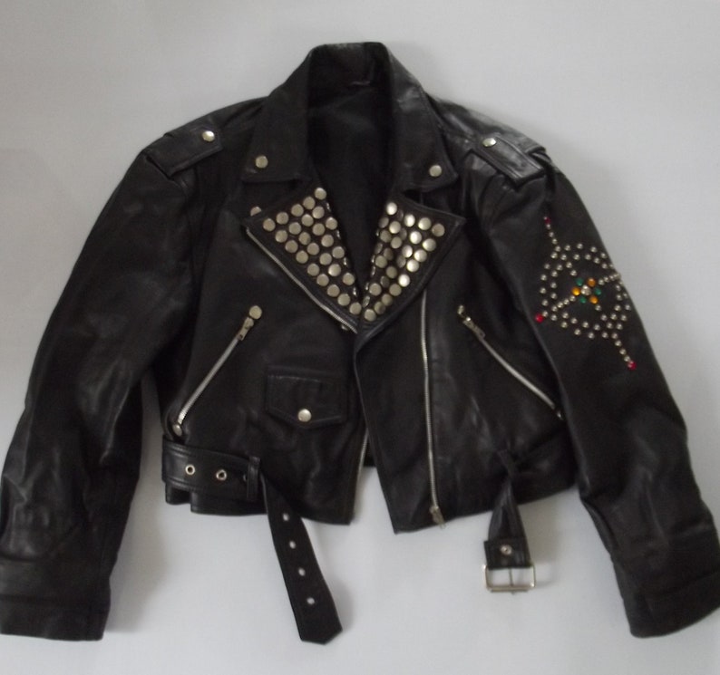 May include: Black leather jacket with silver studs on the collar and shoulder epaulets. The jacket has a diagonal zipper, a belt, and decorative embellishments on the sleeve. A classic biker style.