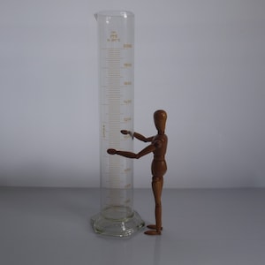 May include: A clear glass graduated cylinder with a capacity of 2000 ml, marked in increments of 100 ml. The cylinder is standing on a white surface. A wooden articulated figure is standing next to the cylinder, with its arms outstretched as if it is holding the cylinder.