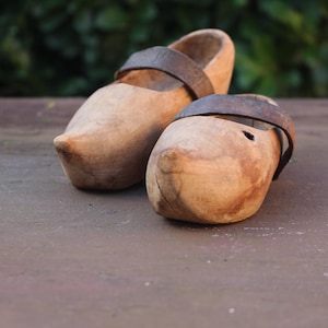 Op de afbeelding: Een paar houten klompen met bruine leren bandjes. De klompen zijn lichtbruin van kleur met een afgeronde neus en een uitgesneden binnenkant. De leren bandjes zijn donkerbruin en bevestigd aan het houten lichaam. Ongeveer 20 cm lang.