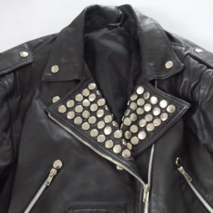 May include: Black leather jacket with silver stud embellishments on the collar and shoulders. Features a zipper closure and multiple silver button details. A classic biker style.