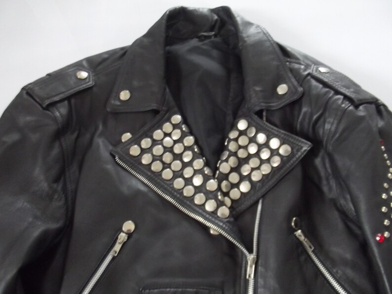 May include: Black leather jacket with silver stud embellishments on the collar and shoulders. Features a zipper closure and multiple silver button details. A classic biker style.