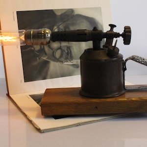Puede incluir: Una lámpara de mesa construida con un soplete de metal antiguo montado sobre una base de madera rectangular. La lámpara cuenta con una bombilla larga de cristal transparente estilo Edison con un filamento brillante en la parte frontal.