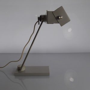 K&ouml;nnte beinhalten: Eine graue Metall-Tischlampe mit einem flexiblen Arm und einem rechteckigen Sockel. Die Lampe hat einen wei&szlig;en Schirm, der verstellt werden kann.
