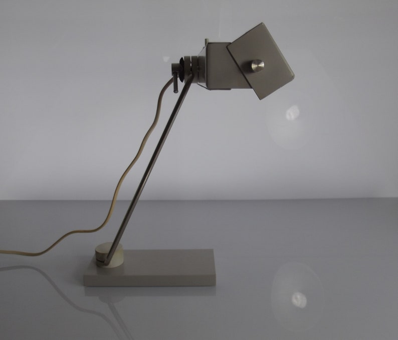 K&ouml;nnte beinhalten: Eine graue Metall-Tischlampe mit einem flexiblen Arm und einem rechteckigen Sockel. Die Lampe hat einen wei&szlig;en Schirm, der verstellt werden kann.
