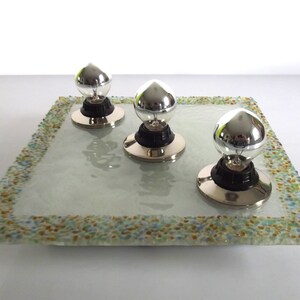 May include: Three silver light bulbs with black bases sit on a square glass tray with a colorful mosaic border.