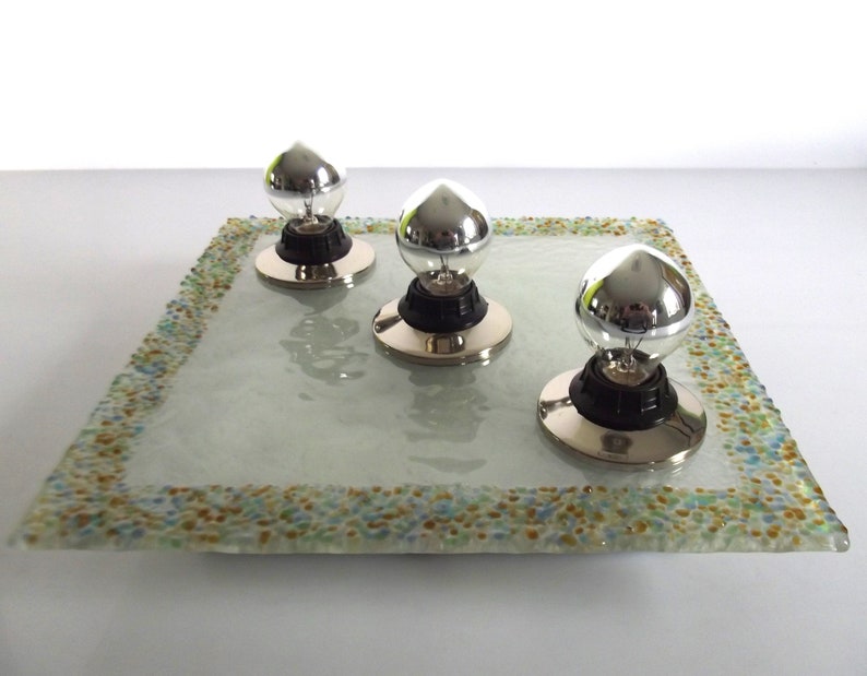 May include: Three silver light bulbs with black bases sit on a square glass tray with a colorful mosaic border.