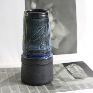 May include: A tall, ceramic vase with a dark blue and black color scheme. The upper portion features a textured leaf design and vertical lines. A dark blue band separates the top from the black base. The vase has a cylindrical shape.